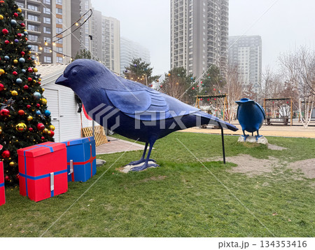 Large blue bird sculpture standing near gift boxes in winter park. Festive urban decor, public art, and holiday atmosphere Large blue bird sculpture standing near gift boxes in winter park. Festive urban decor, public art, and holiday atmosphere 134353416