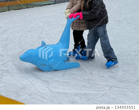 Children learning to skate using a dolphin support on ice. Winter fun, family activity, and beginner skating experience. 134353551