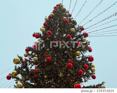 Large outdoor Christmas tree decorated with red and gold balls. Urban winter celebration, public holiday installation and seasonal city decoration 134353624