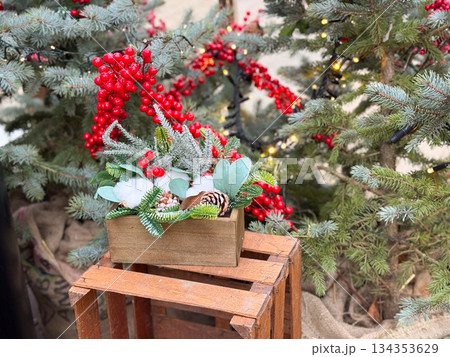 Festive holiday arrangement displayed on wooden crate among fir trees. Christmas market decor, seasonal styling and winter atmosphere Festive holiday arrangement displayed on wooden crate among fir trees. Christmas market decor, seasonal styling and winter atmosphere 134353629