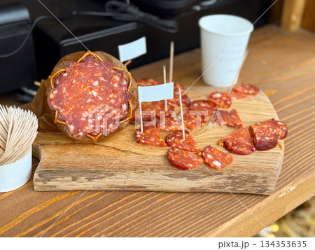 Sliced cured sausage served on wooden board at market stall. Street food presentation, traditional snack and rustic food styling 134353635
