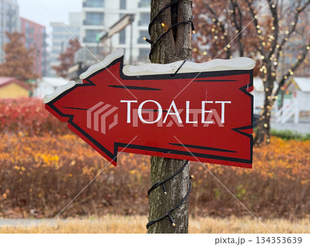 Red directional sign reading toilet attached to tree in festive urban park. Winter holiday navigation detail, seasonal event infrastructure and humorous street element. Red directional sign reading toilet attached to tree in festive urban park. Winter holiday navigation detail, seasonal event infrastructure and humorous street element. 134353639