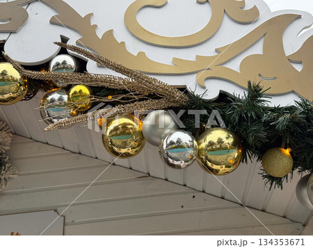 Golden and silver Christmas ornaments arranged under decorative sign on festive wooden stall roof. Holiday market decor, winter celebration atmosphere and traditional seasonal design. Golden and silver Christmas ornaments arranged under decorative sign on festive wooden stall roof. Holiday market decor, winter celebration atmosphere and traditional seasonal design. 134353671