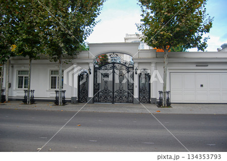 Ornate black gate of a grand white residence. Ornate black wrought iron gate stands as the centerpiece of a grand white residence, featuring intricate scrollwork and decorative patterns. 134353793