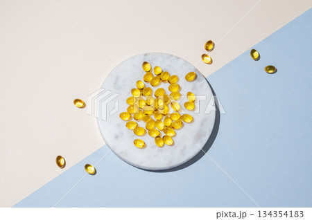 Yellow softgel vitamins capsules on a white marble plate on a blue background with shadow. Yellow softgel vitamins capsules on a white marble plate on a blue background with shadow. 134354183