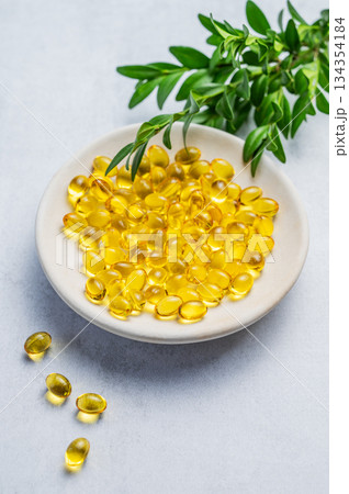 Yellow softgel vitamins capsules on a white plate with green leaves on a blue background Yellow softgel vitamins capsules on a white plate with green leaves on a blue background 134354184