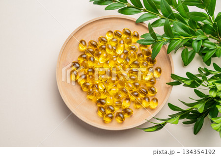Yellow softgel vitamins capsules in a wooden plate on a light background with green leaf. Yellow softgel vitamins capsules in a wooden plate on a light background with green leaf. 134354192