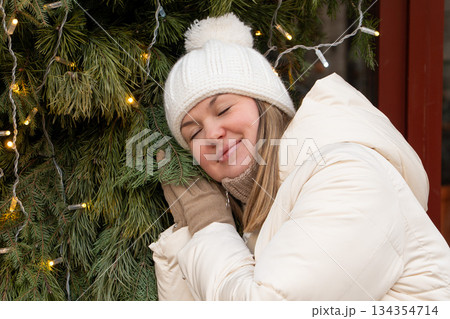 Smiling woman in a white winter jacket and knitted hat leans against a decorated evergreen with warm fairy lights, creating a calm, cozy holiday mood 134354714
