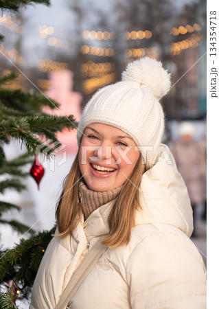 Smiling young woman in a white winter coat and knitted hat stands by a decorated Christmas tree with warm lights on Tverskoy Boulevard in Moscow Smiling young woman in a white winter coat and knitted hat stands by a decorated Christmas tree with warm lights on Tverskoy Boulevard in Moscow 134354718
