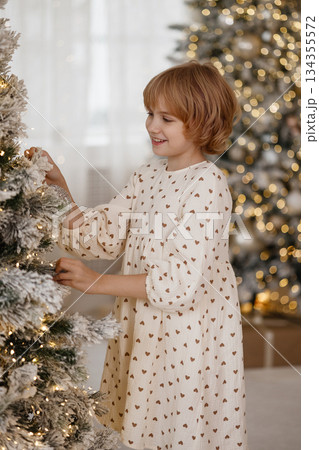 Side view of Caucasian blonde girl decorating Christmas tree in cozy room 134355572
