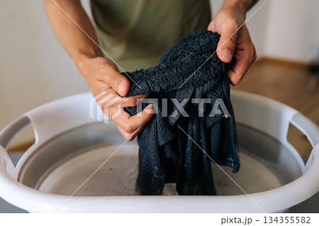 Handwashing dark clothing in plastic basin, wringing wet garment with bare hands over gray surface, showcasing manual laundry technique and water conservation. Concept of hygiene and cleanliness. 134355582