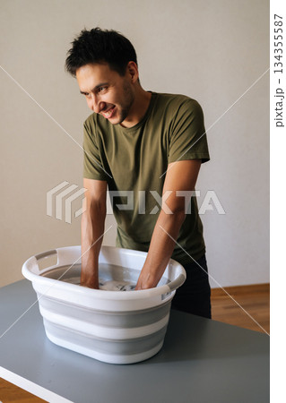 Vertical portrait of positive young man washing clothes by hand in plastic basin, wringing out water performing domestic task, smiling looking away. Concept of housework, housekeeping and household. 134355587