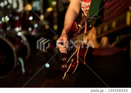 Guitarist Plays Energetically, Warm Amber Lights Highlight Guitarist Strumming Over Drums In Background 134356420