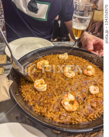 Close-up of Spanish paella with shrimp and metal spoon on table beside glass of beer. Authentic Mediterranean cuisine, dining, and travel concept. High quality photo Close-up of Spanish paella with shrimp and metal spoon on table beside glass of beer. Authentic Mediterranean cuisine, dining, and travel concept. High quality photo 134358970