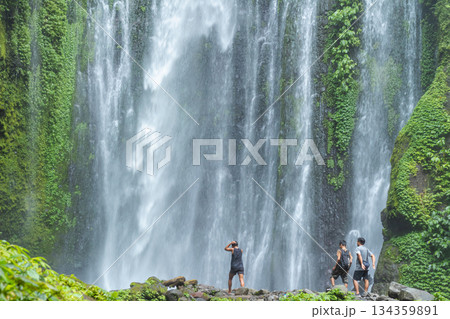 LOMBOK UTARA, INDONESIA - JANUARY 27, 2025 - Majestic tropical waterfall cascades down a moss-covered cliff face, with travelers exploring the rocky pool edge in Lombok, Southeast Asia. LOMBOK UTARA, INDONESIA - JANUARY 27, 2025 - Majestic tropical waterfall cascades down a moss-covered cliff face, with travelers exploring the rocky pool edge in Lombok, Southeast Asia. 134359891