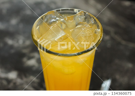Macro shot of bright orange tropical refreshment drink with melting ice cubes in a tall glass, set against a blurred dark background. 134360075