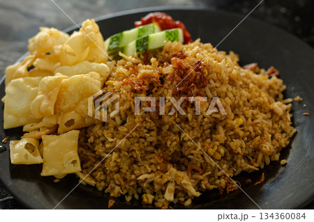 Authentic Indonesian Nasi Goreng served hot, garnished with fried shallots, shredded chicken, alongside crispy pangsit crackers, fresh cucumber, and spicy chili paste. 134360084