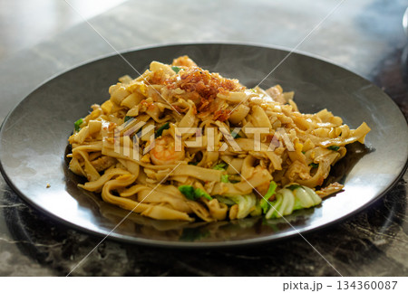 High angle close-up of steaming Kwetiau Goreng, a savory Indonesian stir-fried flat noodle dish featuring shrimp, egg, and cabbage, garnished with crispy fried shallots. 134360087