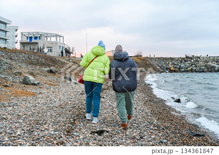 Two Russian friends aged 50 and 60 walk along the cold spring shore, talking and sharing calm connection in warm clothes with the sea beside them. 134361847
