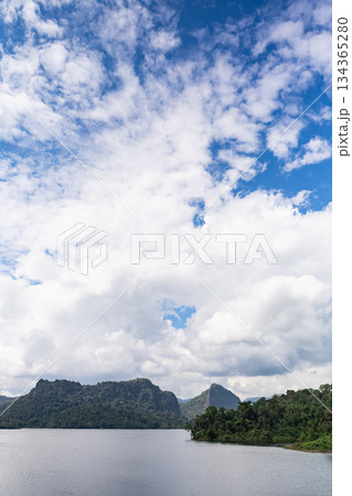 Serene Landscape of Calm Water Surrounded by Lush Green Hills Under a Bright Blue Sky with Fluffy White Clouds in the Distance 134365280