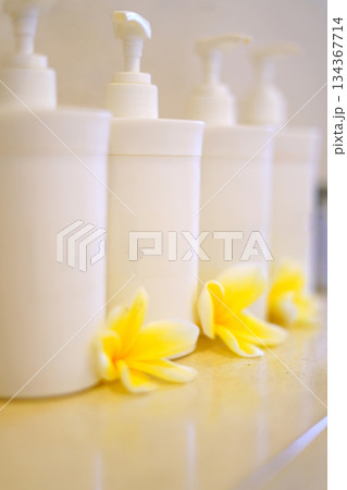 Soft focus image featuring white pump bottles arranged in a row with fresh yellow plumeria flowers at the base. Clean, spa-like scene ideal for beauty, skincare, and wellness branding. 134367714