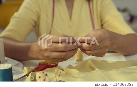 Asian woman threading the needle. Tailor working in her studio with focus in hands putting thread in sewing needle. Asian woman threading the needle. Tailor working in her studio with focus in hands putting thread in sewing needle. 134368358