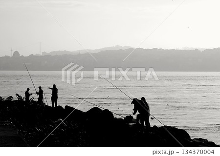Silhouettes of Bosphorus fishermen in the early morning. A black-and-white, high-contrast image that captures the serene solitude of recreational fishing in Istanbul. Silhouettes of Bosphorus fishermen in the early morning. A black-and-white, high-contrast image that captures the serene solitude of recreational fishing in Istanbul. 134370004