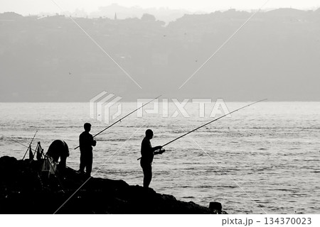 Silhouettes of Bosphorus fishermen in the early morning. A black-and-white, high-contrast image that captures the serene solitude of recreational fishing in Istanbul. 134370023