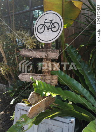 A wooden bicycle sign in a garden, with arrows indicating directions. Green plants surround the sign, creating a natural scene 134373428
