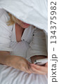 Smiling woman lying on a bed with a blanket over her head while using the mobile phone 134375982