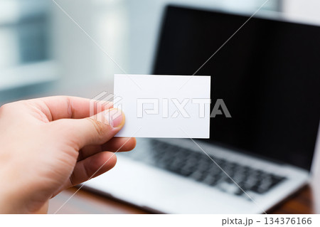 Business card in hand on laptop background. Hand holding blank business card in front of laptop. Blank white business card mockup in office environment. Business card in hand on laptop background. Hand holding blank business card in front of laptop. Blank white business card mockup in office environment. 134376166