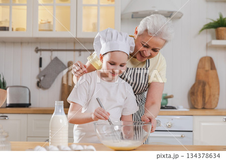 Happy family in kitchen. Grandmother and granddaughter child cook in kitchen together. Grandma teaching kid girl knead dough bake cookies. Household teamwork helping family generations concept Happy family in kitchen. Grandmother and granddaughter child cook in kitchen together. Grandma teaching kid girl knead dough bake cookies. Household teamwork helping family generations concept 134378634