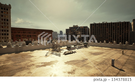 Bright sunlight casts an artistic glow on a deserted rooftop. Old debris lies across the surface, surrounded by towering buildings under a dramatic sky. The scene captures urban solitude. 134378815