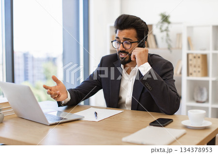 An Arabian businessman wearing glasses is working on his laptop while talking on the phone in his office An Arabian businessman wearing glasses is working on his laptop while talking on the phone in his office 134378953
