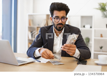 An Indian businessman is carefully counting a stack of US dollar bills at his desk, possibly for financial planning or investment purposes An Indian businessman is carefully counting a stack of US dollar bills at his desk, possibly for financial planning or investment purposes 134378964