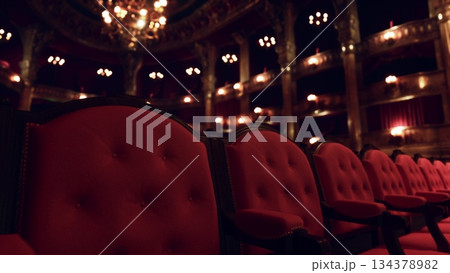 Rows of plush red chairs fill the beautiful theater, softly illuminated by ornate chandeliers. The atmosphere is inviting, stirring excitement for the upcoming performance. 134378982