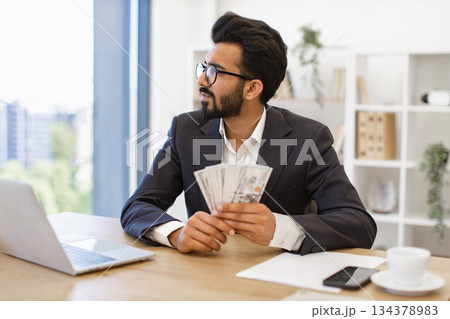 An Indian businessman in a suit holds a stack of cash while looking out the window 134378983