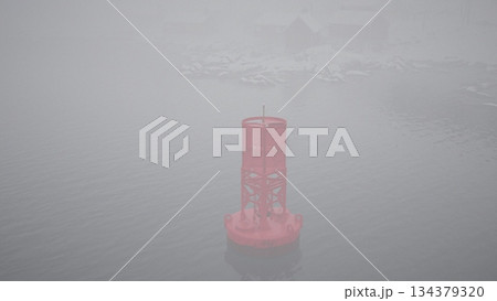 A vivid pink buoy rises from the calm, misty waters, surrounded by dense fog. Snowfall lightly dusts the scene, evoking a serene, quiet atmosphere in a coastal location. A vivid pink buoy rises from the calm, misty waters, surrounded by dense fog. Snowfall lightly dusts the scene, evoking a serene, quiet atmosphere in a coastal location. 134379320