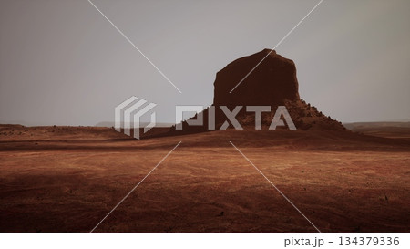 A striking rock formation rises from a barren terrain, surrounded by endless stretches of reddish soil and an atmosphere that hints at isolation. The scene evokes a sense of wonder and exploration. 134379336