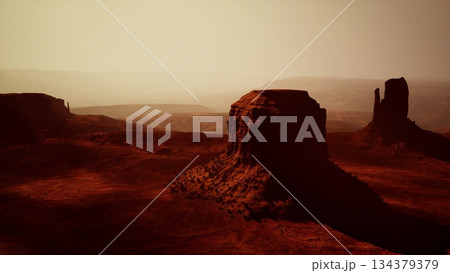 At dusk, warm hues illuminate the vast desert landscape of Monument Valley, highlighting towering red rock formations. Shadows stretch across the sandy terrain, creating a serene atmosphere. 134379379