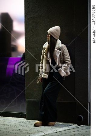 Fashionable young woman in cozy winter attire leans against a stylish city wall during twilight hours, capturing the essence of urban chic 134379569