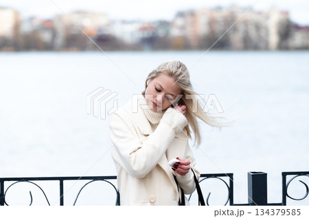 Beautiful young woman with flowing hair stands by a tranquil lake, reflecting on life amidst cool breezes and a picturesque urban backdrop in early spring Beautiful young woman with flowing hair stands by a tranquil lake, reflecting on life amidst cool breezes and a picturesque urban backdrop in early spring 134379585