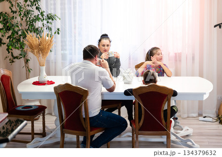 Gathering of family at a large dining table enjoying warm beverages and sharing stories during a cozy afternoon in a sunlit room filled with plants Gathering of family at a large dining table enjoying warm beverages and sharing stories during a cozy afternoon in a sunlit room filled with plants 134379632
