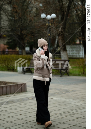 Crisp autumn day in the park with a young woman talking on her phone while dressed warmly in a stylish coat and knit hat Crisp autumn day in the park with a young woman talking on her phone while dressed warmly in a stylish coat and knit hat 134379638