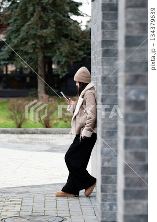 Fashionable woman using smartphone while leaning against a stone wall in a modern urban setting with greenery in the background during a chilly afternoon Fashionable woman using smartphone while leaning against a stone wall in a modern urban setting with greenery in the background during a chilly afternoon 134379639
