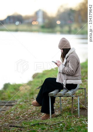 Enjoying a peaceful moment by the riverside while absorbed in a mobile device during a chilly evening in autumn Enjoying a peaceful moment by the riverside while absorbed in a mobile device during a chilly evening in autumn 134379642