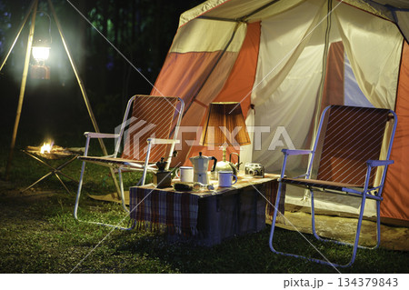 Camping tents at night, vintage cabin tents, antique oil lamps, retro chairs, and a group of camping tents with outdoor coffee-making facilities on wooden tables in a forest camping area. Camping tents at night, vintage cabin tents, antique oil lamps, retro chairs, and a group of camping tents with outdoor coffee-making facilities on wooden tables in a forest camping area. 134379843