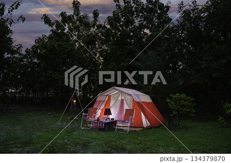 Camping tents at night, vintage cabin tents, antique oil lamps, retro chairs, and a group of camping tents with outdoor coffee-making facilities on wooden tables in a forest camping area. Camping tents at night, vintage cabin tents, antique oil lamps, retro chairs, and a group of camping tents with outdoor coffee-making facilities on wooden tables in a forest camping area. 134379870