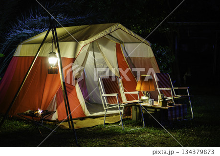 Camping tents at night, vintage cabin tents, antique oil lamps, retro chairs, and a group of camping tents with outdoor coffee-making facilities on wooden tables in a forest camping area. Camping tents at night, vintage cabin tents, antique oil lamps, retro chairs, and a group of camping tents with outdoor coffee-making facilities on wooden tables in a forest camping area. 134379873
