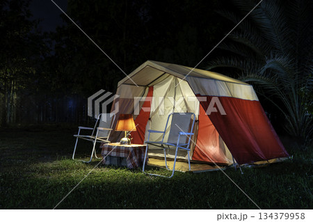 Camping tents at night, vintage cabin tents, antique oil lamps, retro chairs, and a group of camping tents with outdoor coffee-making facilities on wooden tables in a forest camping area. Camping tents at night, vintage cabin tents, antique oil lamps, retro chairs, and a group of camping tents with outdoor coffee-making facilities on wooden tables in a forest camping area. 134379958